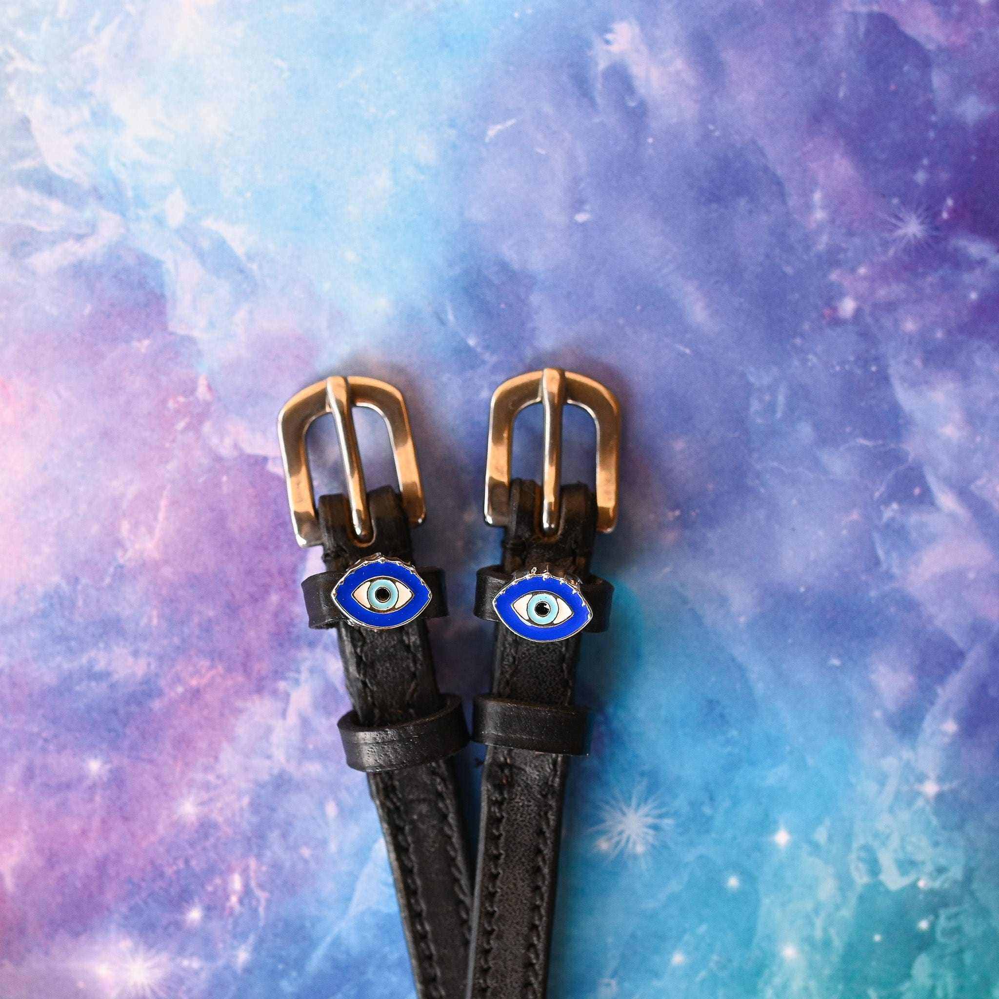 Black leather evil eye spur straps with brass buckles on a galaxy print background