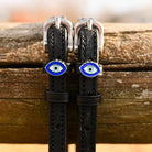 Close-up of blue evil eye charms on black leather spur straps over a wooden rail