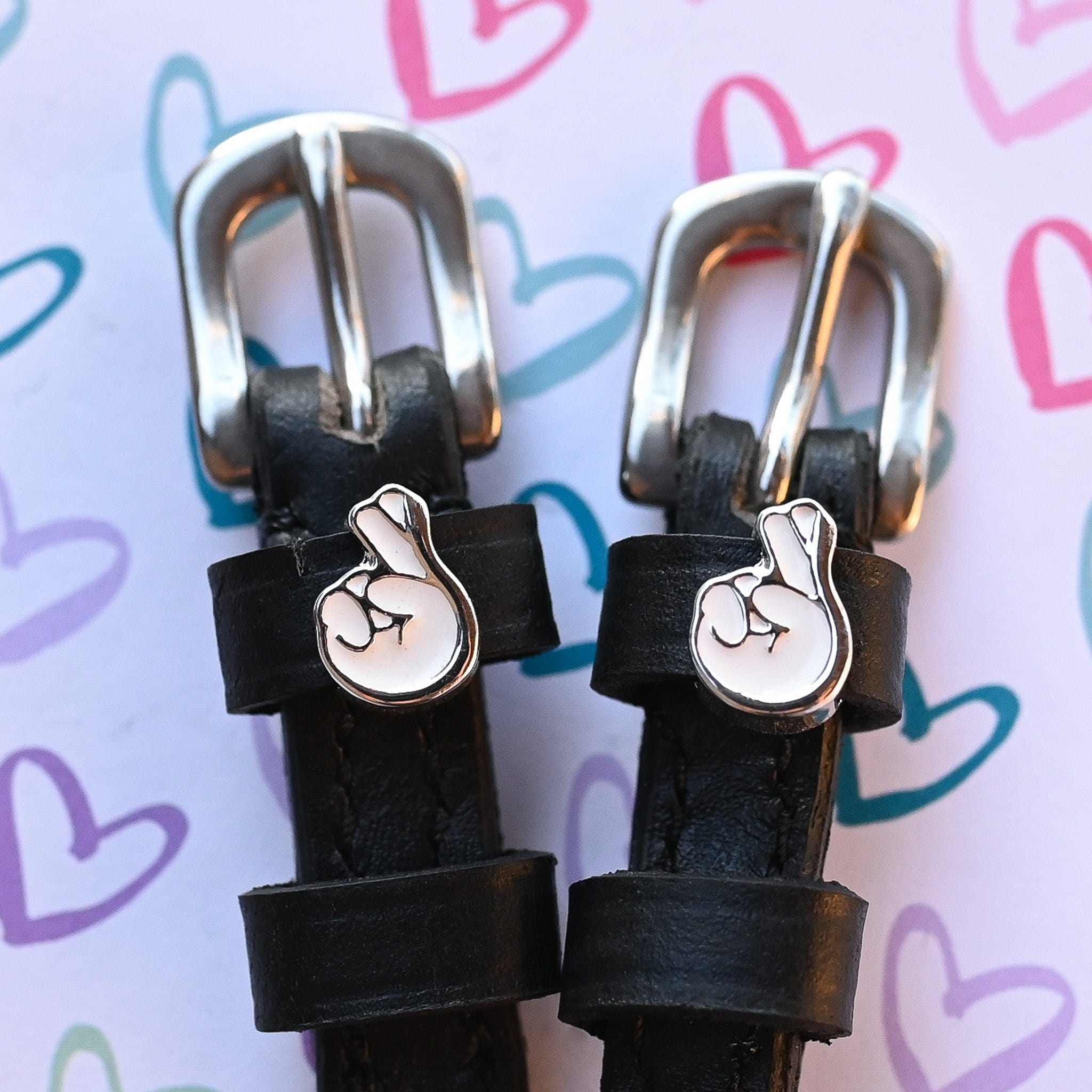 Silver crossed-fingers charms on black spur strap keepers against a hand-drawn hearts print