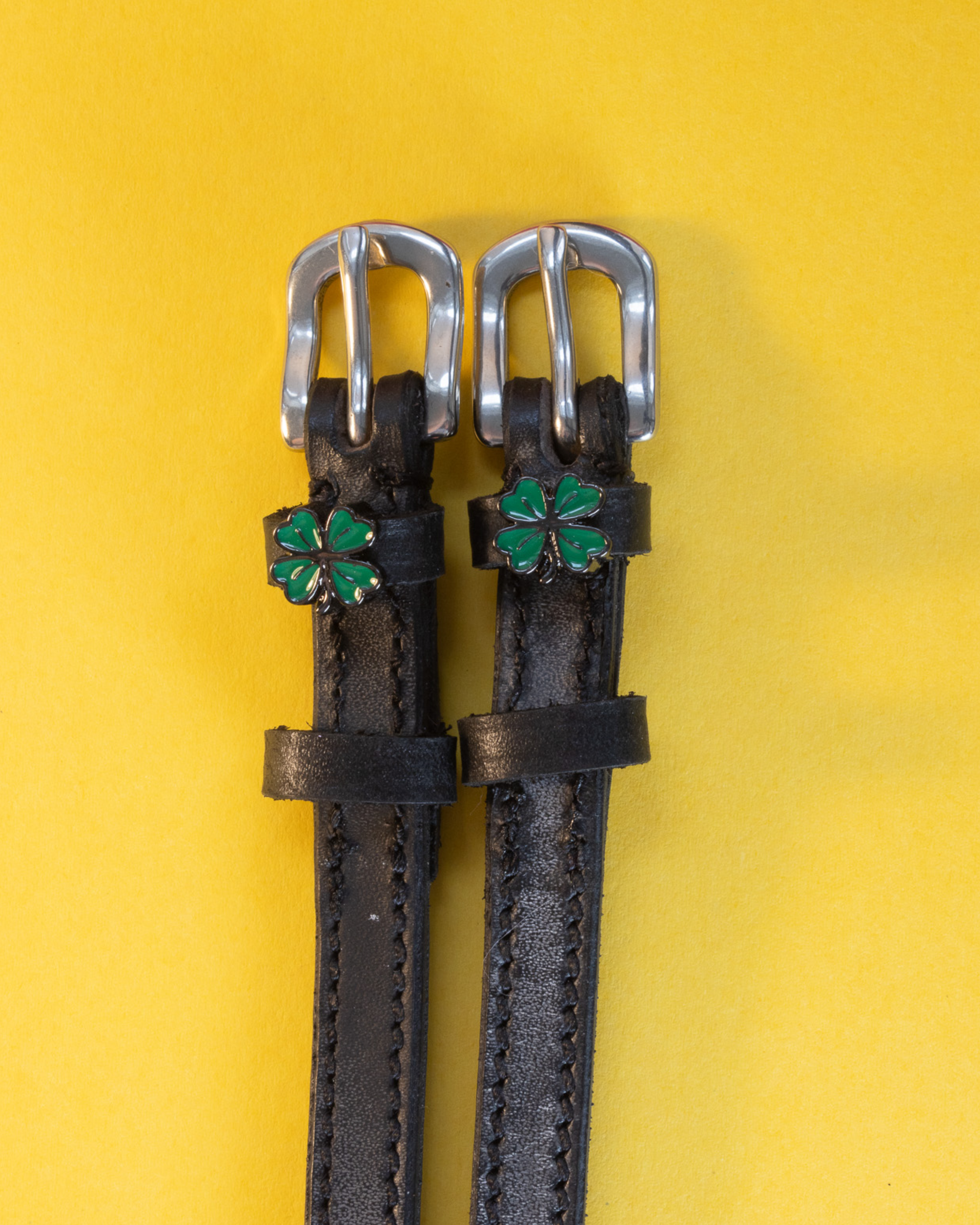 Black leather ManeJane spur straps with green four leaf clover charms on a yellow background