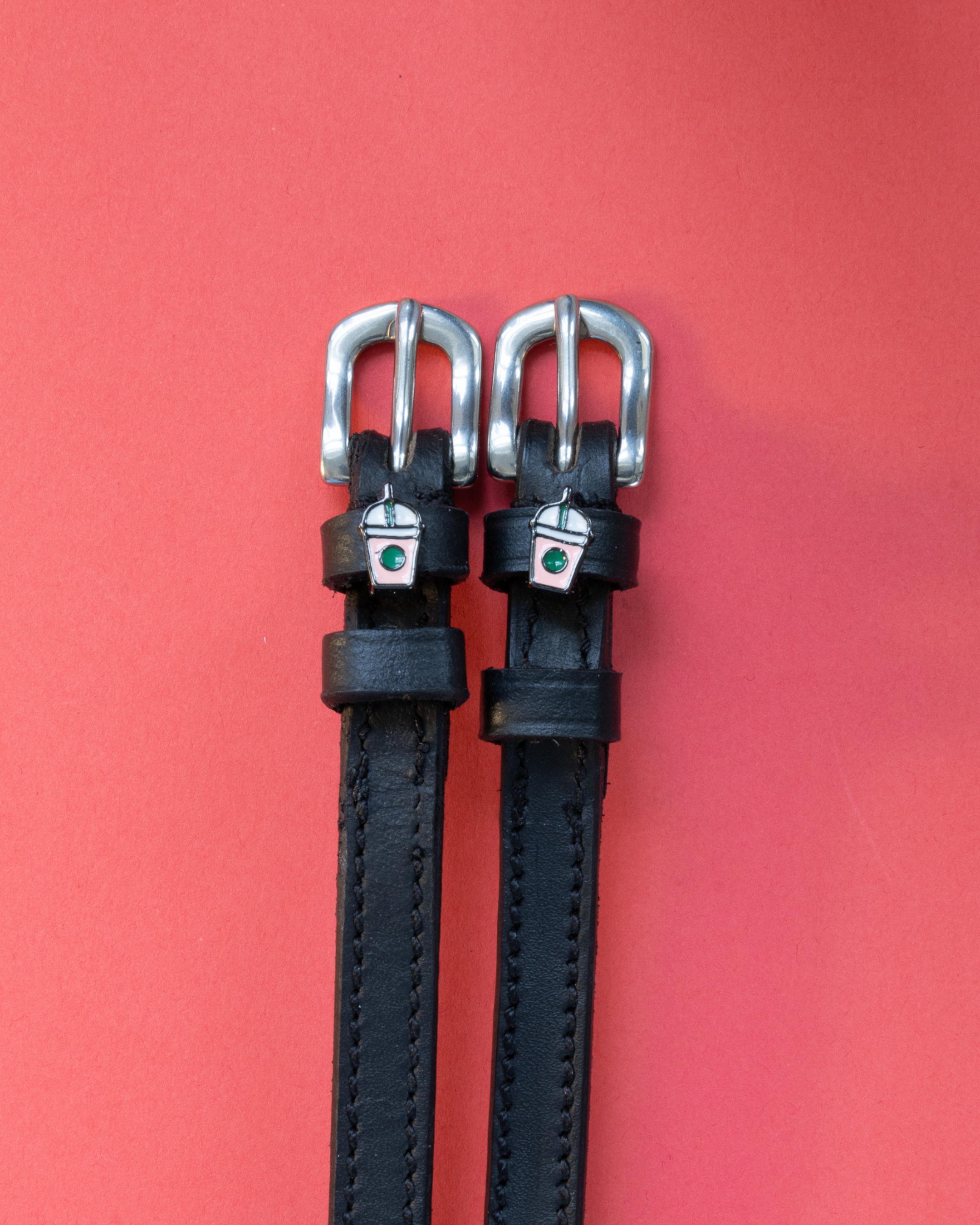 Black leather ManeJane spur straps with silver frappe cup charms on a red background