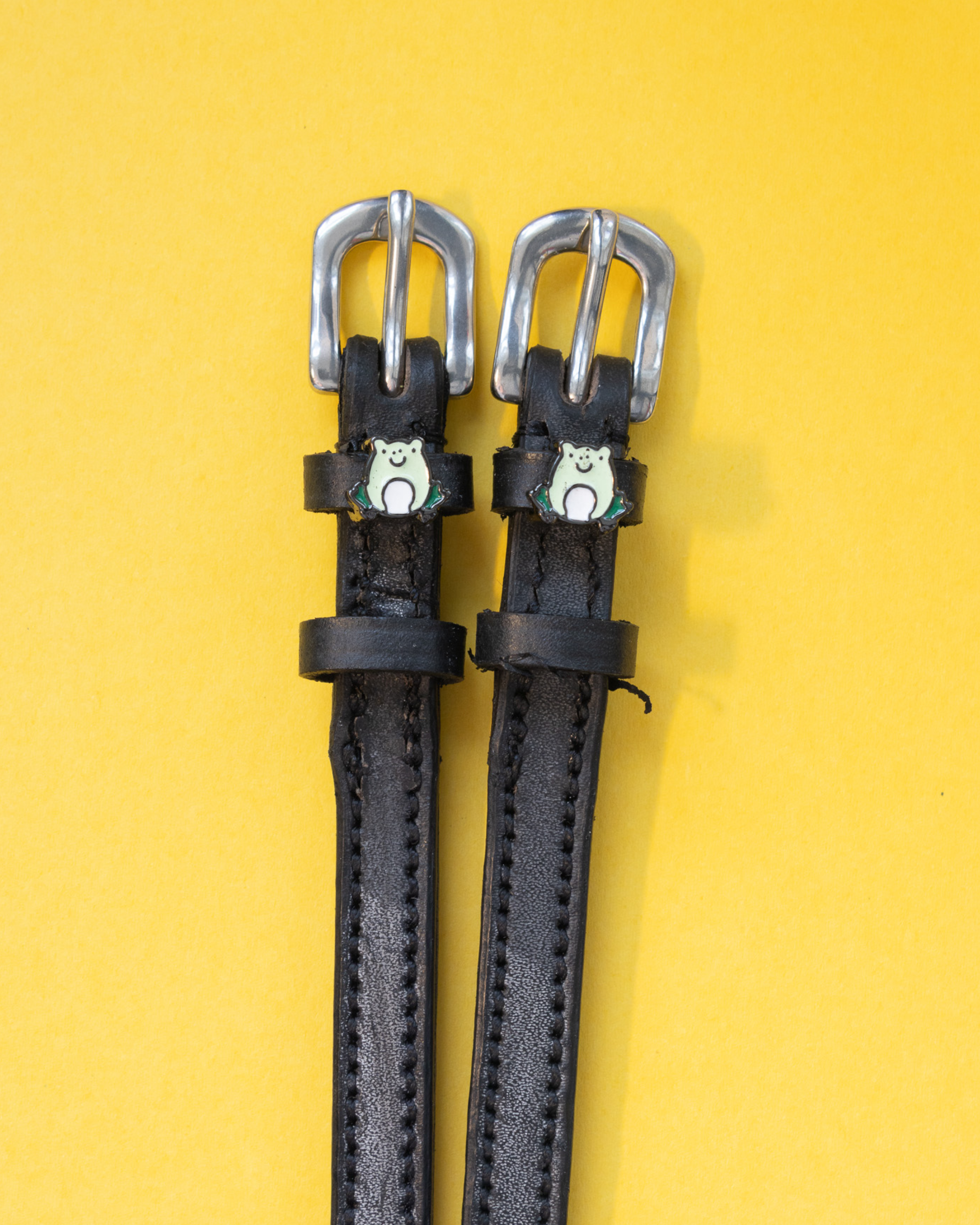 Black leather ManeJane spur straps with silver buckles and green frog charms on a yellow background