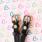 Black leather frog spur straps with brass buckles on a pastel heart-patterned background