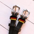 German flag charm spur straps in black leather on a pale pink wood-plank background