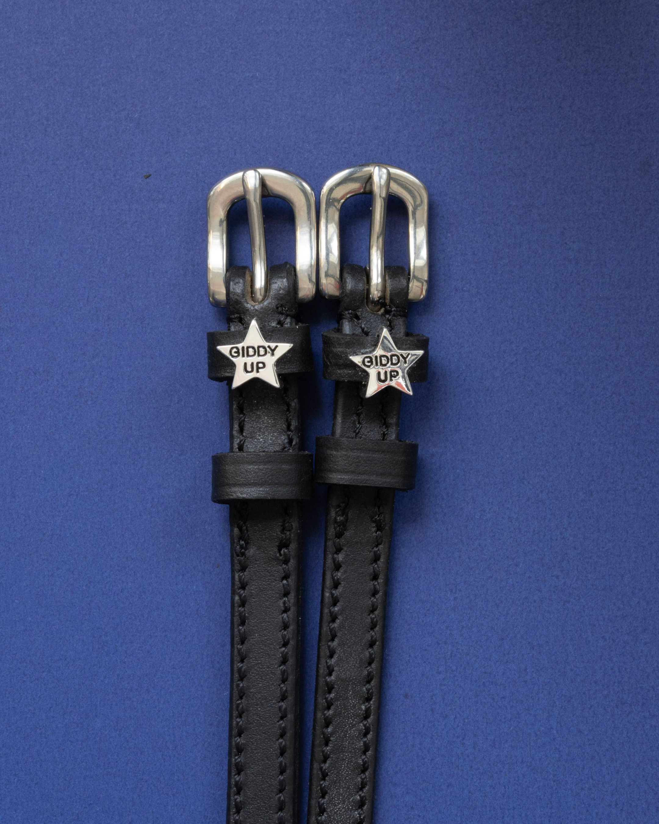 Black leather ManeJane spur straps with silver 'Giddy Up' star charms on a blue background
