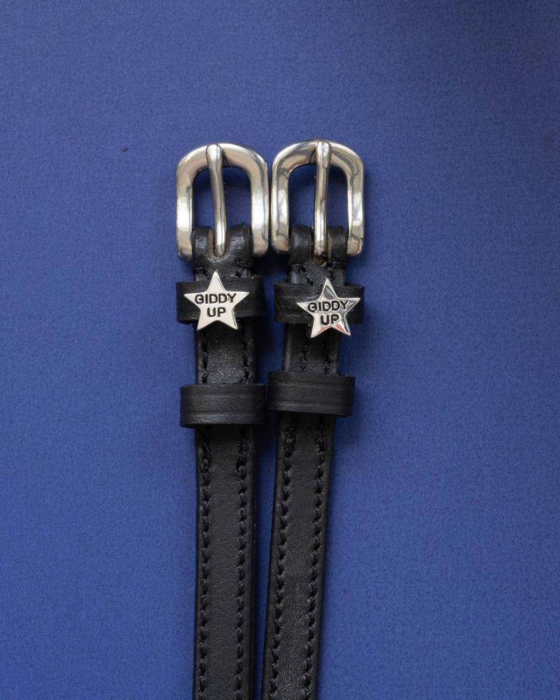 Black leather ManeJane spur straps with silver 'Giddy Up' star charms on a blue background