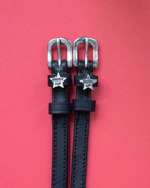 Black leather ManeJane 'Giddy Up' star spur straps with silver buckles on a coral background