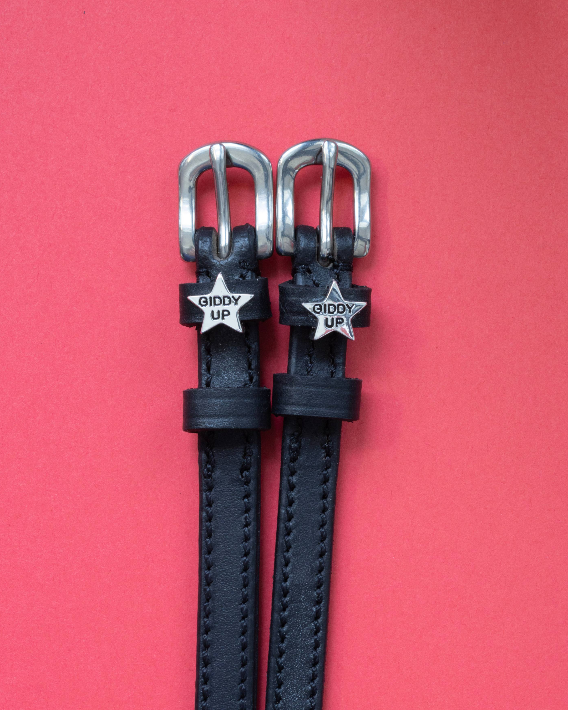 Black leather ManeJane 'Giddy Up' star spur straps with silver buckles on a coral background