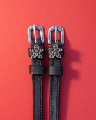 Black leather ManeJane spur straps with gingerbread man charms on a red background