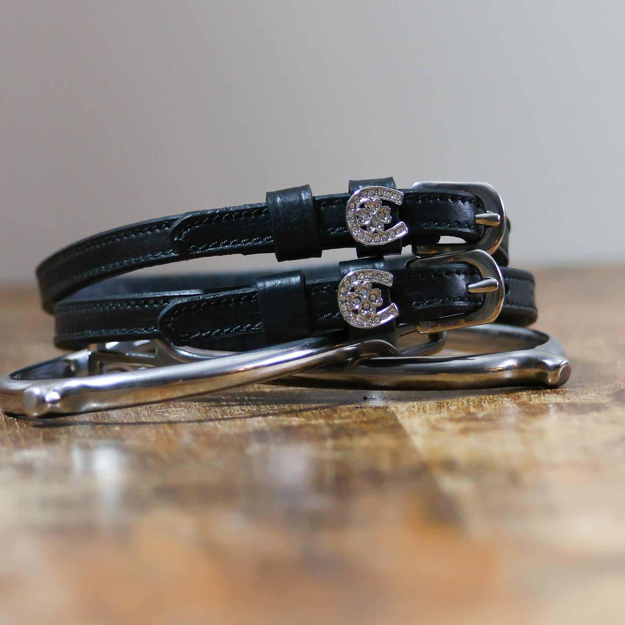 Coiled black leather spur straps with sparkling horseshoe-clover charms stacked on a spur