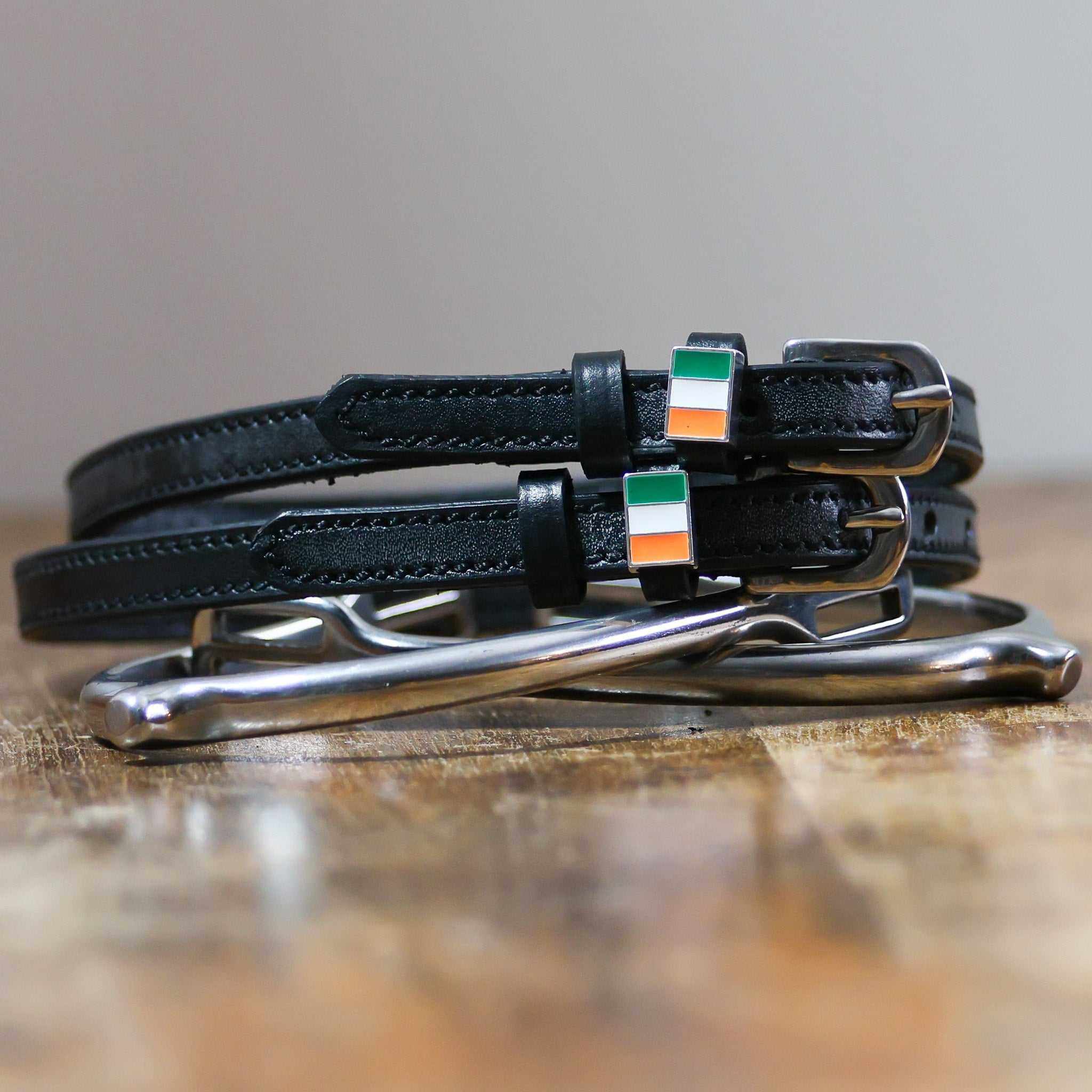 Black leather Ireland flag spur straps coiled beside a stirrup on wood
