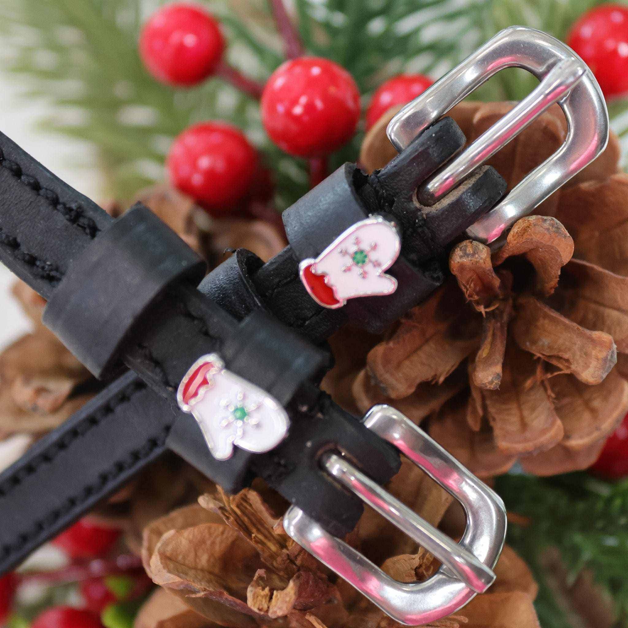 Close-up of white winter mitten charms on black leather spur straps among pine cones and berries