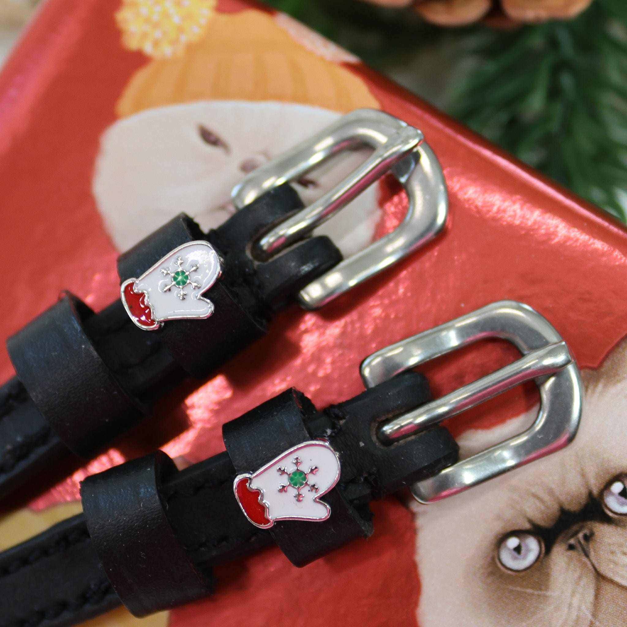 Angled close-up of white mitten charms on black leather spur straps against a red holiday box