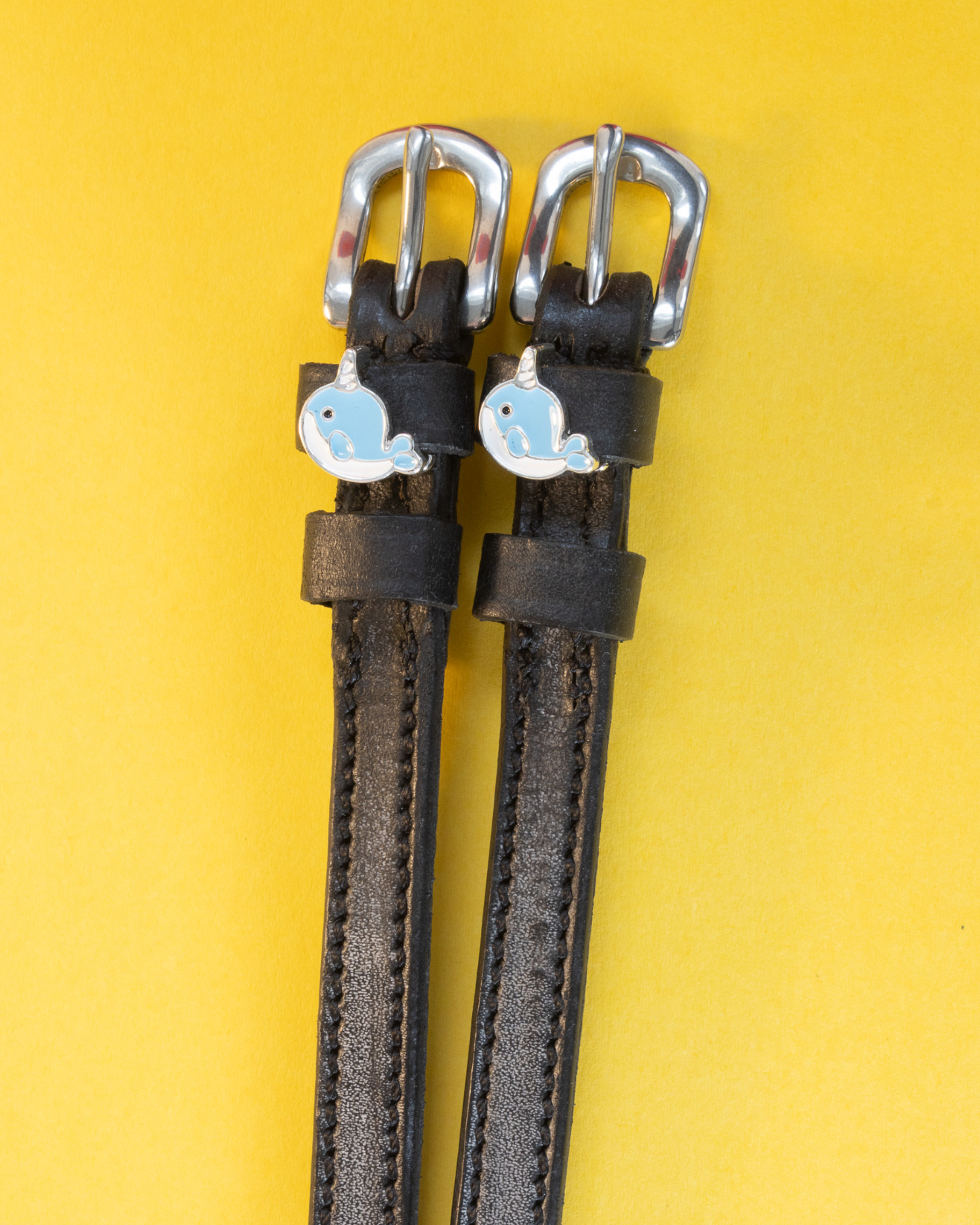 Black leather ManeJane spur straps with blue enamel narwhal charms on a yellow background