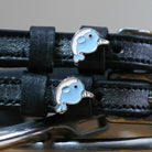 Close-up of two blue enamel narwhal charms on black leather ManeJane spur strap keepers