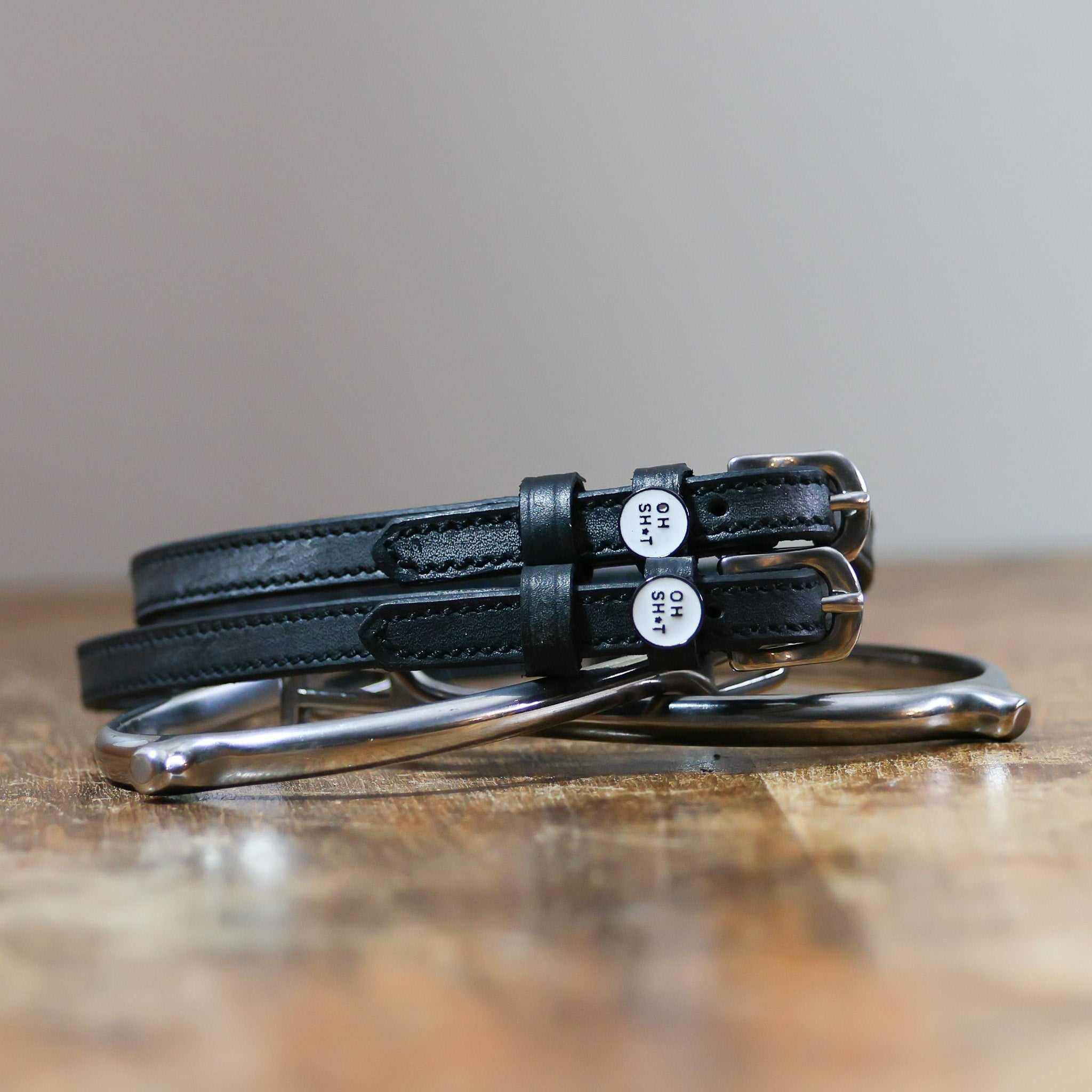 Black leather OH SH*T spur straps coiled beside a stirrup on wood