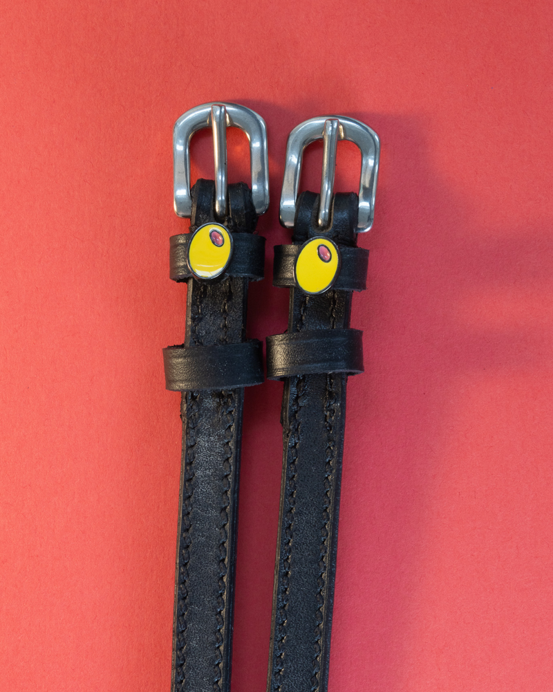 Black leather ManeJane spur straps with green olive charms on a coral red background