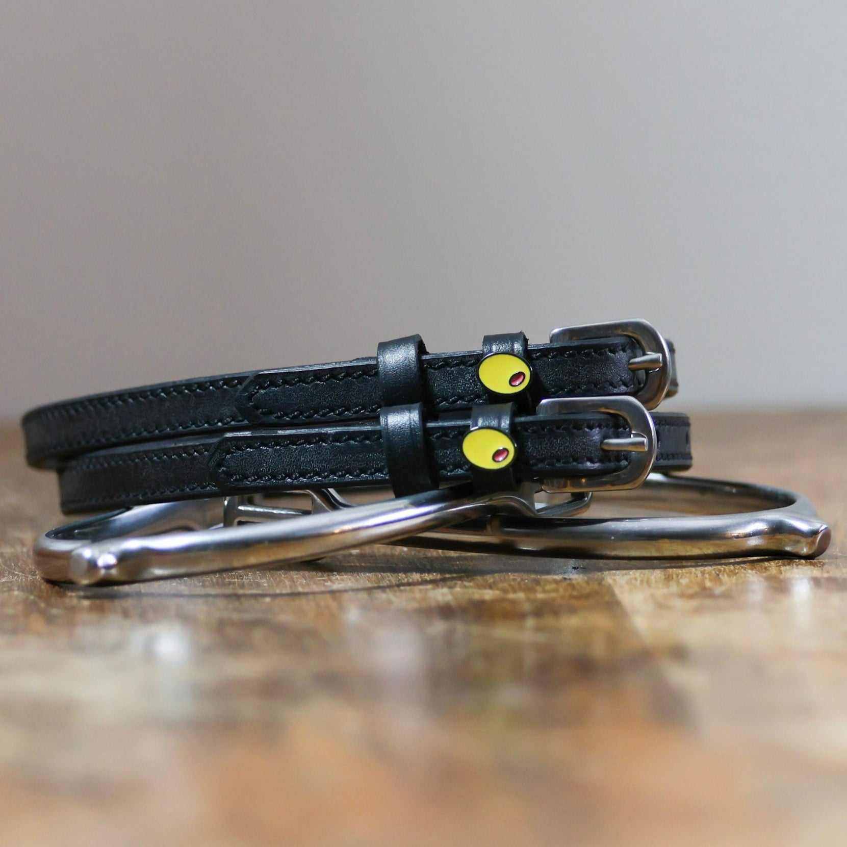 Black leather olive spur straps coiled on a wooden surface with a silver bit