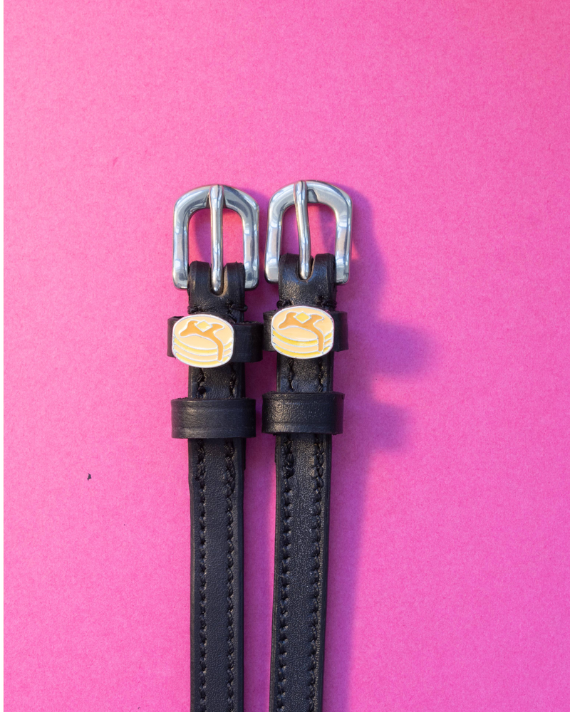 Black leather ManeJane pancake spur straps with silver buckles, flat-laid on a pink background