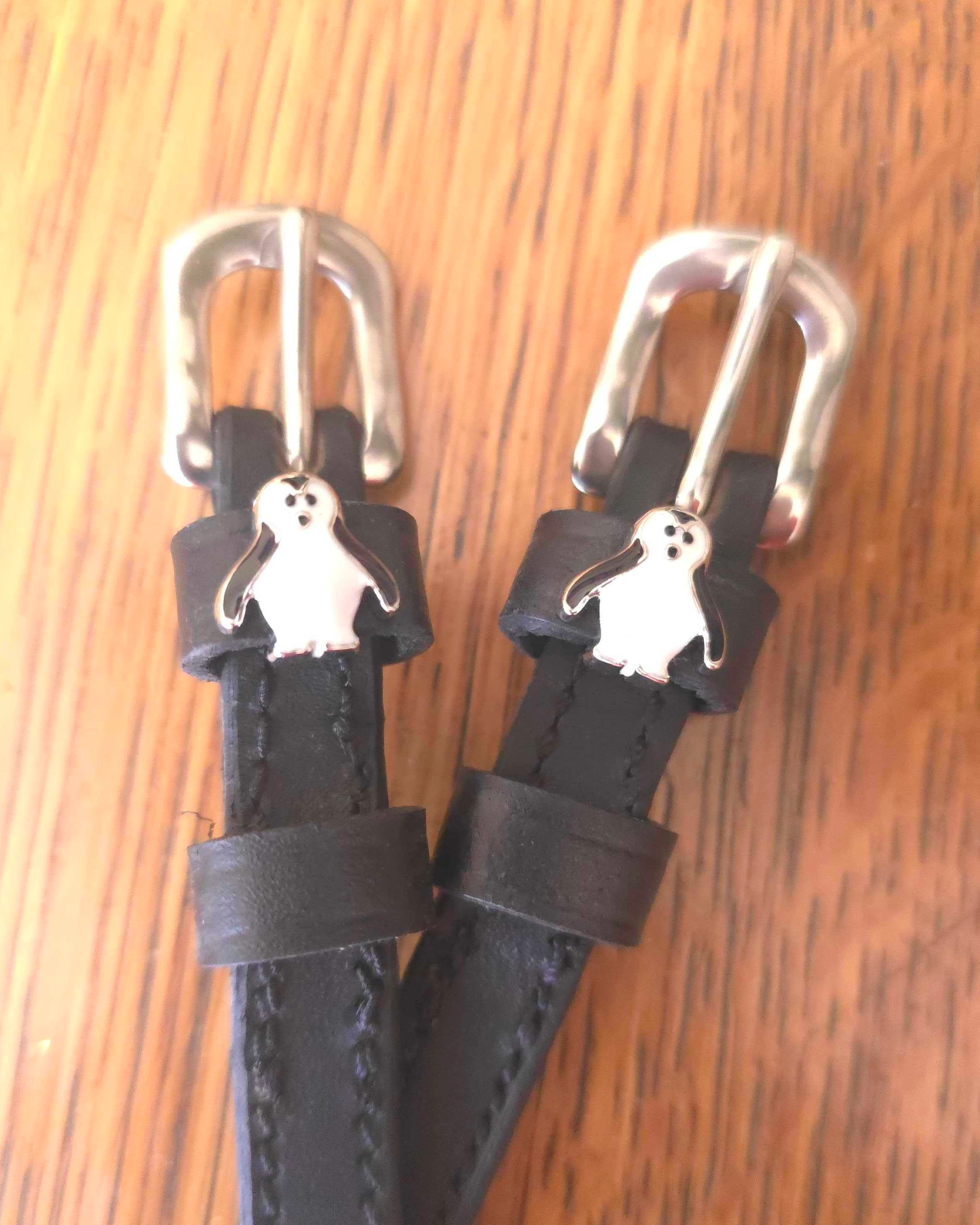 Close-up of silver penguin charms on black leather ManeJane spur straps over wood grain