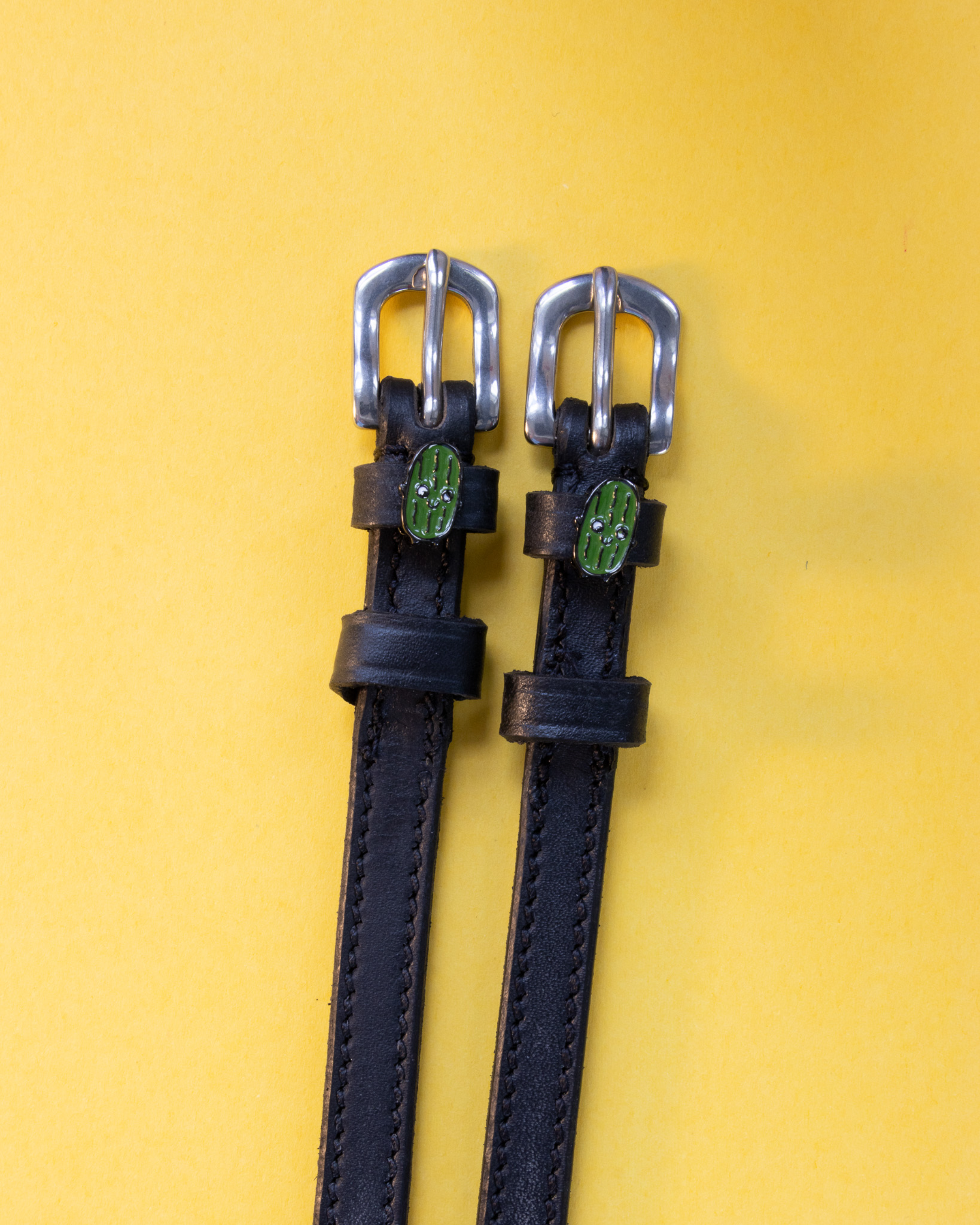 Pair of black leather pickle spur straps with silver buckles laid flat on yellow