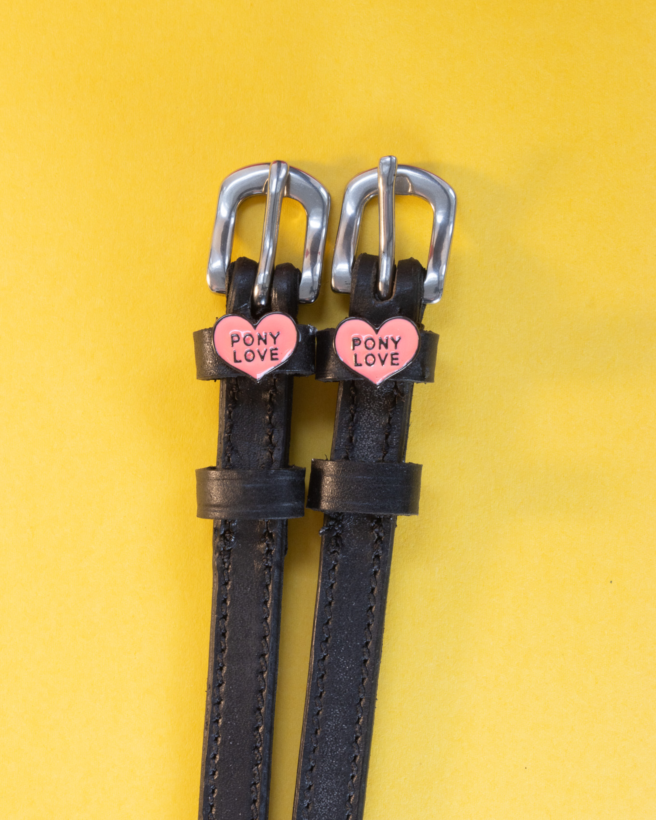 Black leather Pony Love spur straps with silver buckles on a yellow background
