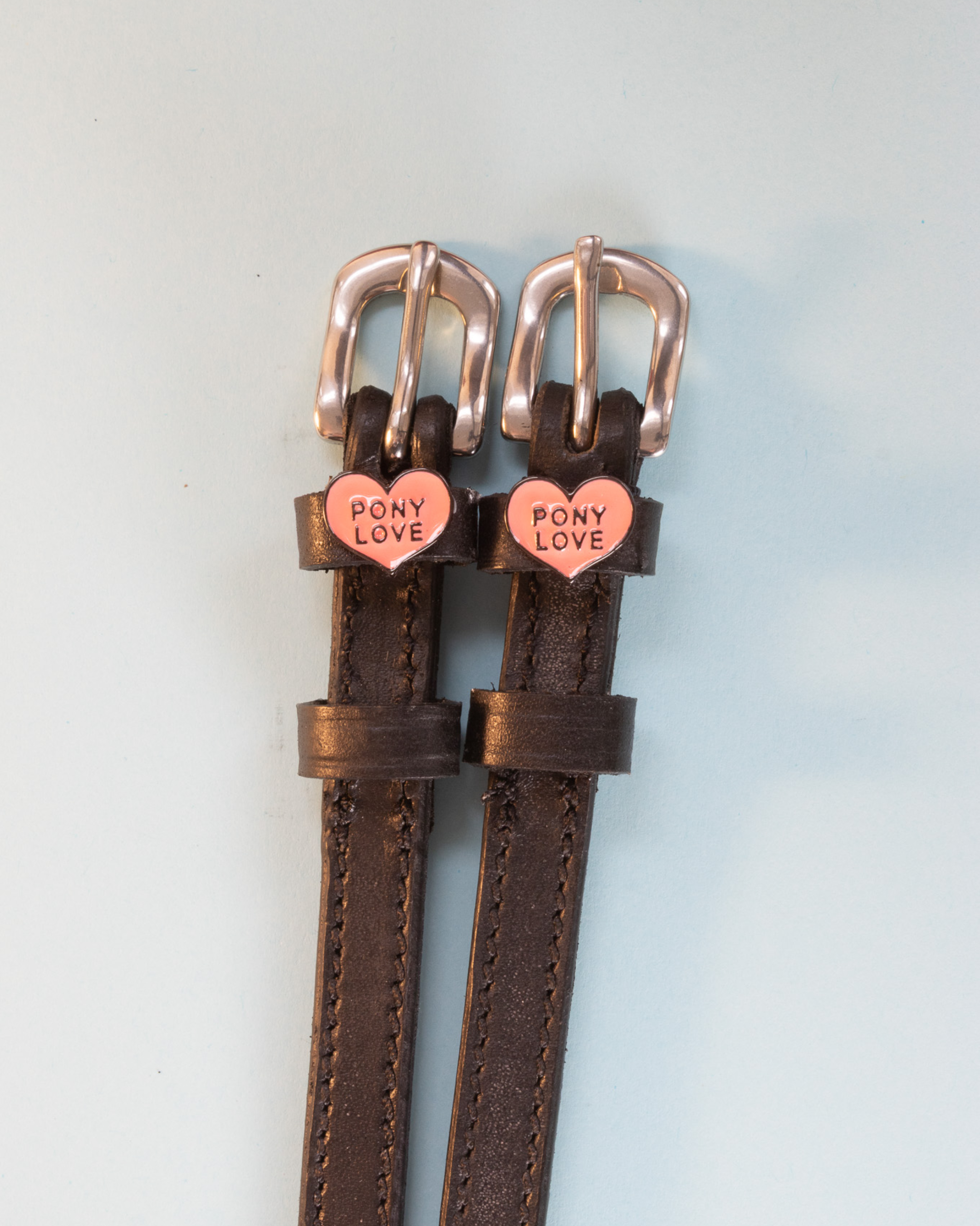 Brown leather ManeJane Pony Love spur straps on a soft blue-grey background