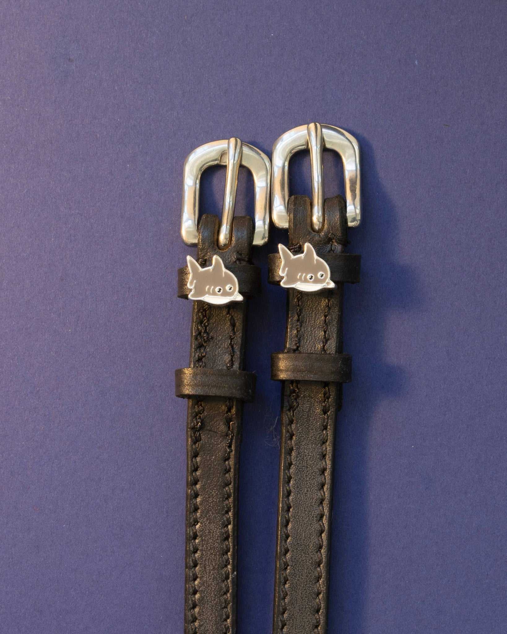 Silver shark charm spur straps on black leather against a deep purple background