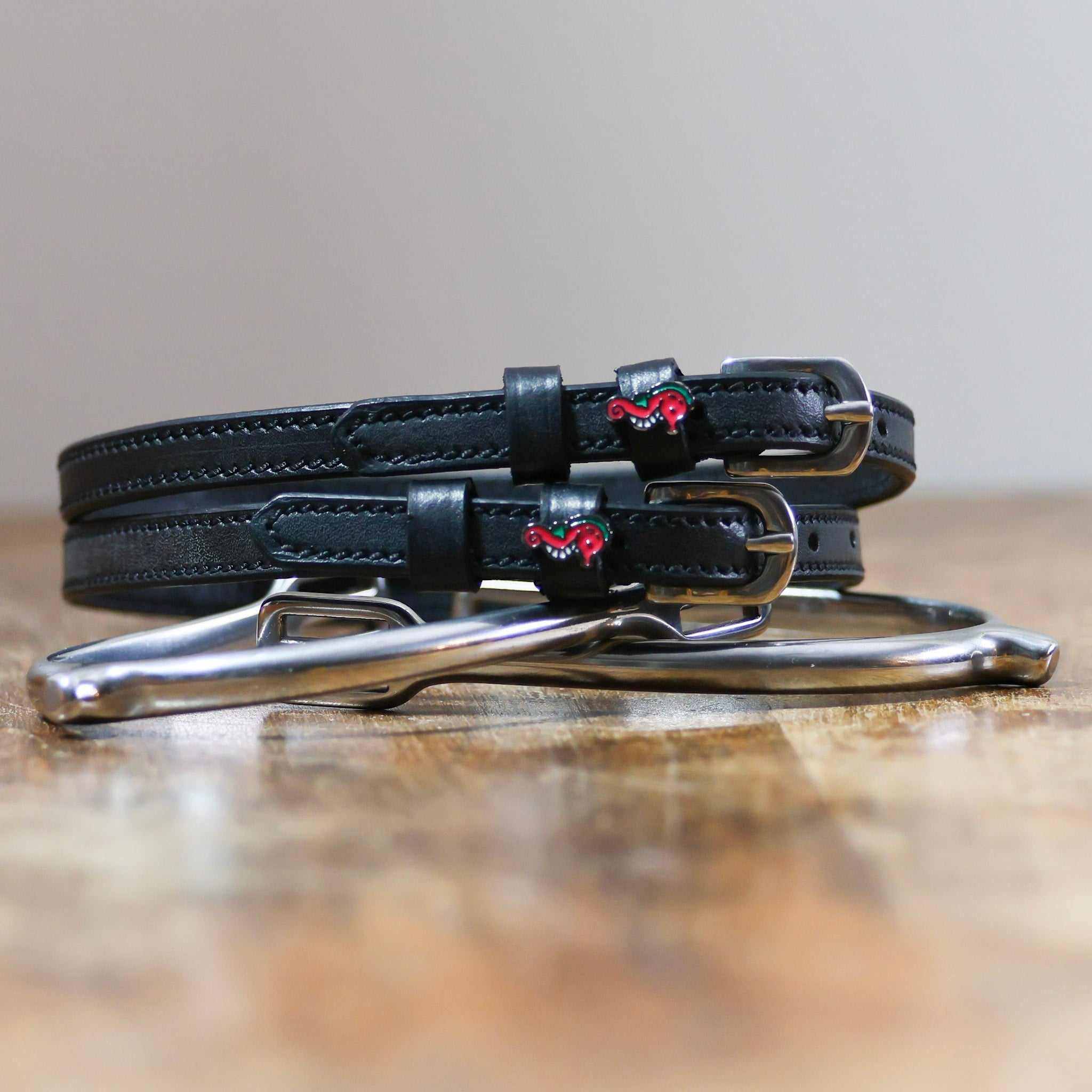 Black leather seahorse spur straps coiled beside a stirrup on a wooden surface