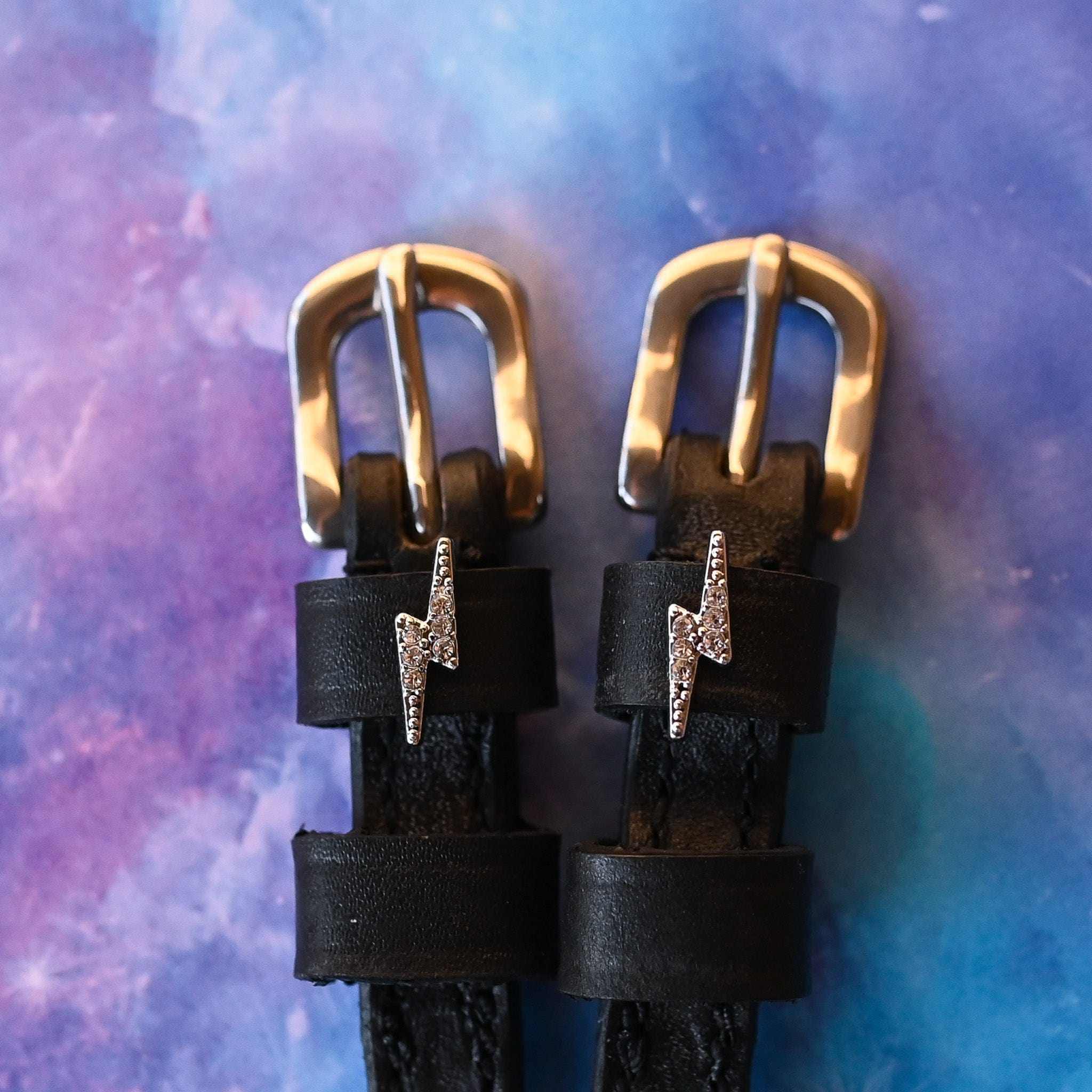 Close-up of rhinestone lightning bolt charms on black spur straps over a galaxy backdrop