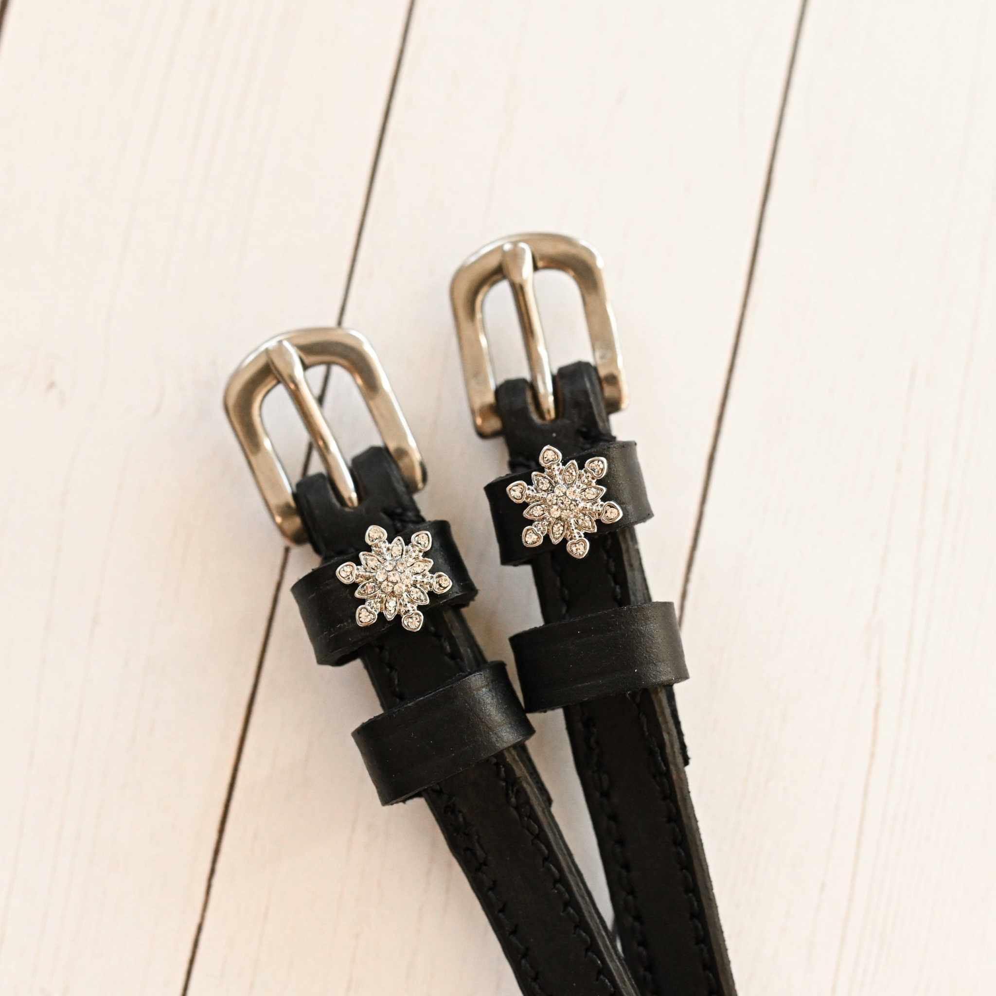 Close-up of black snowflake spur straps with crystal charms on white-washed wood planks