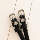 Close-up of black snowflake spur straps with crystal charms on white-washed wood planks