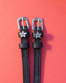 Black leather spur straps with silver 'The Boss' star charms laid flat on a coral background