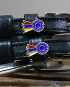Close-up of tri-color ribbon rosette charms on black leather spur straps beside a bit