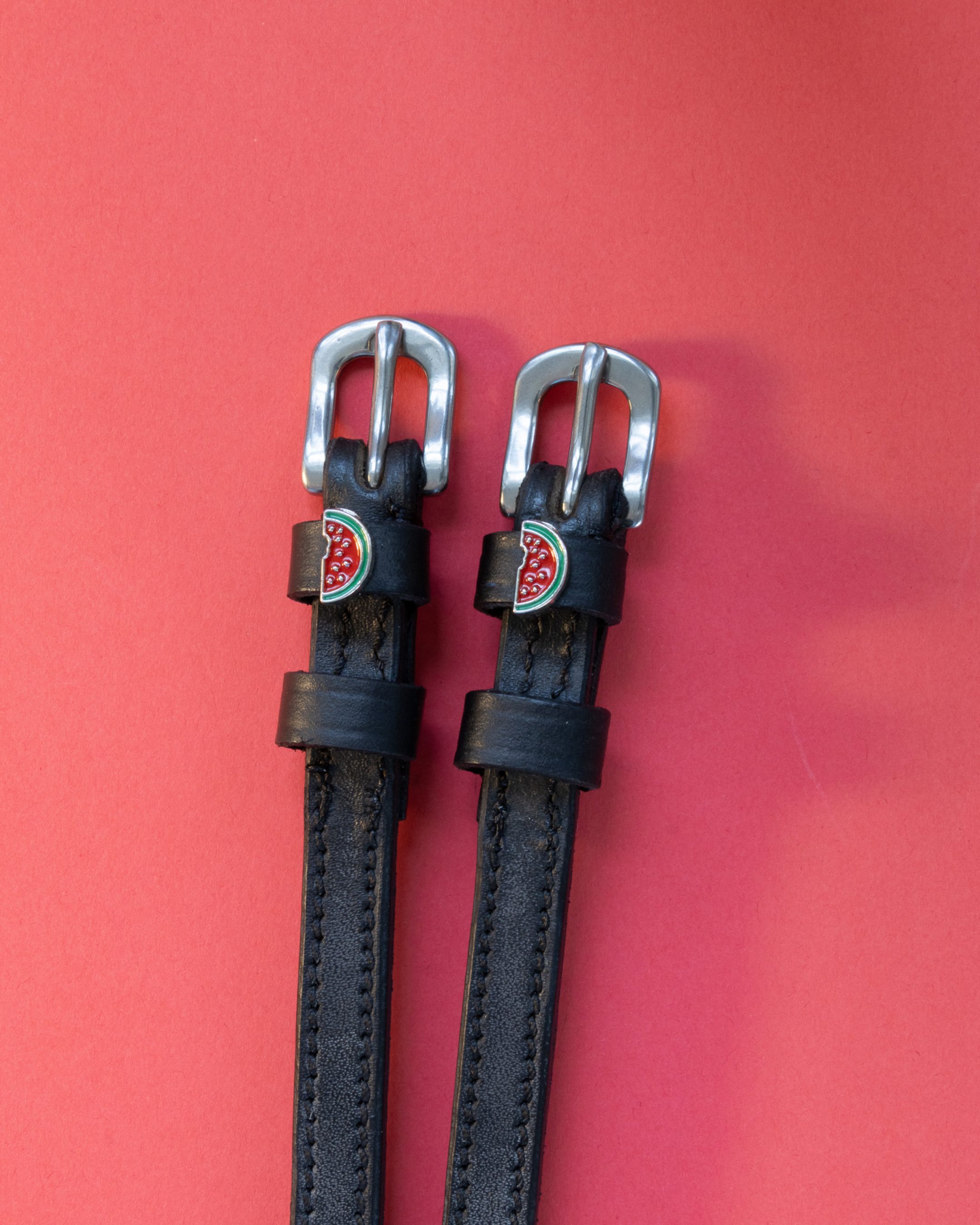 Black leather ManeJane spur straps with silver buckles and enamel watermelon charms on red background