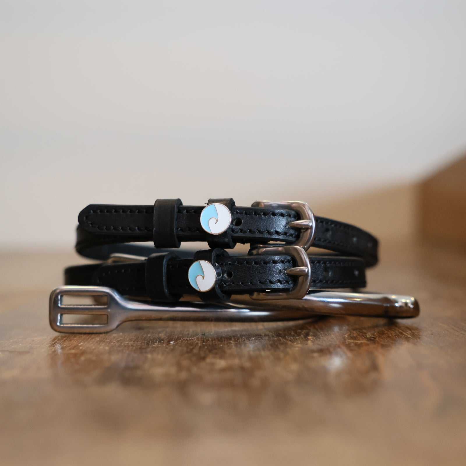 Black leather wave spur straps coiled on a wooden surface with a silver bit