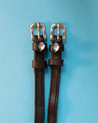 Black leather ManeJane shark charm spur straps with silver buckles on a light blue background
