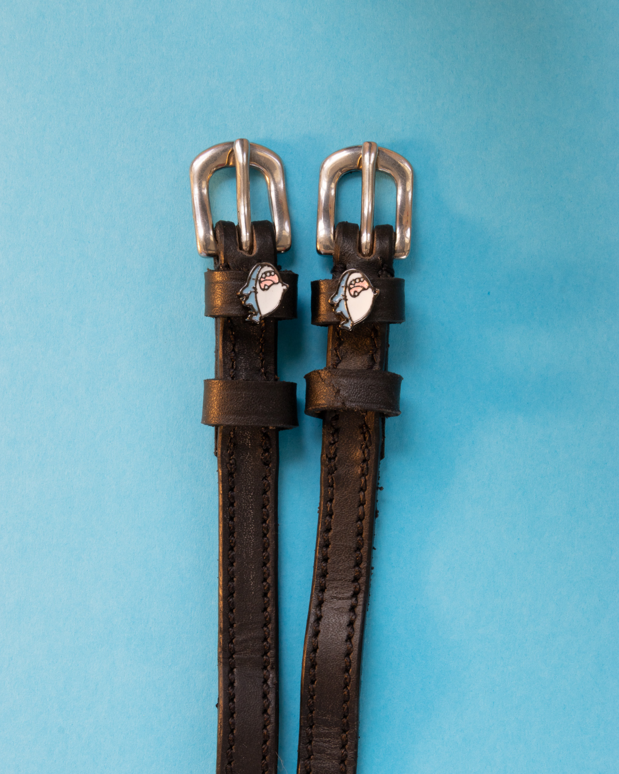 Black leather ManeJane shark charm spur straps with silver buckles on a light blue background