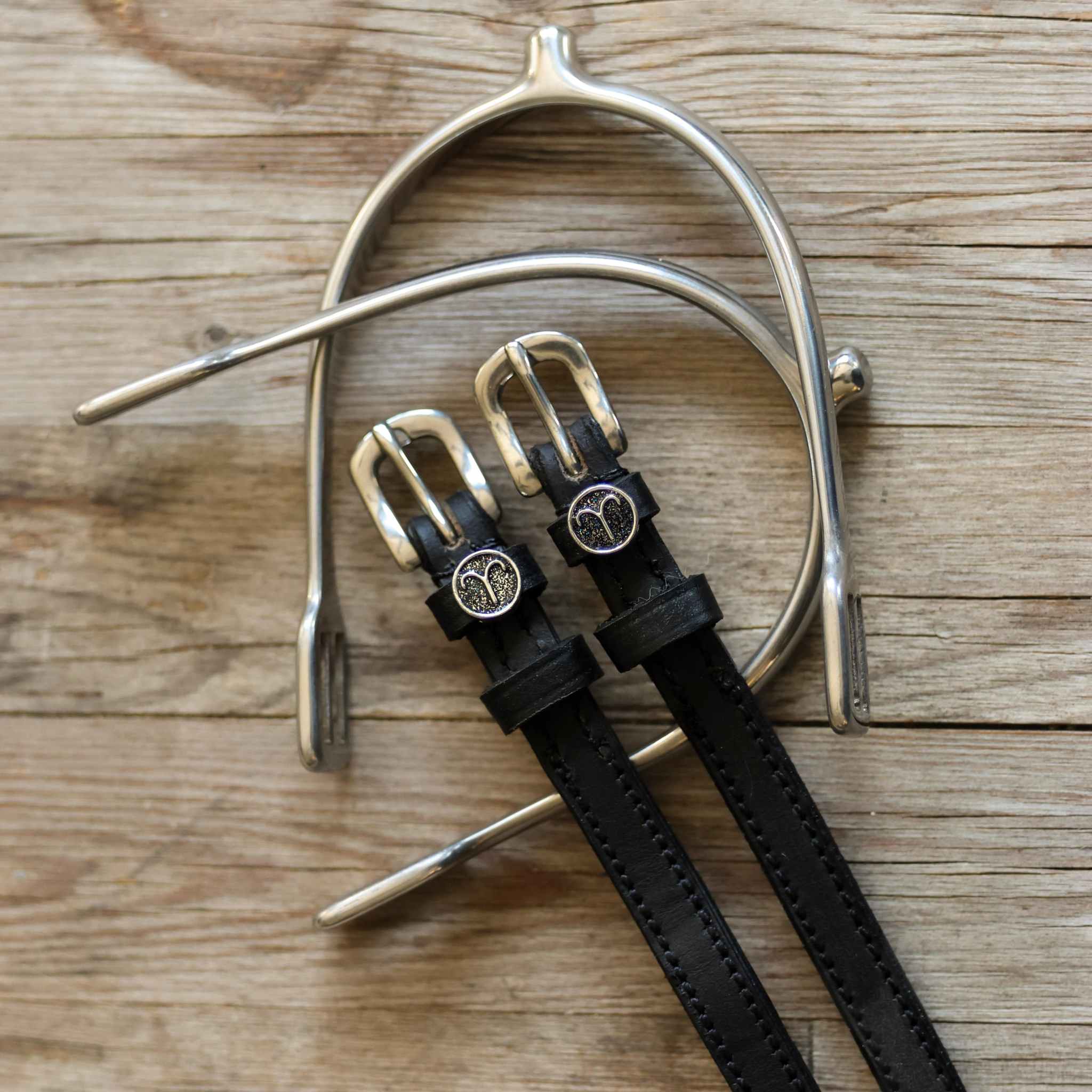 Black leather Aries Spur Straps with stainless steel buckles and custom Aries charm on wooden background