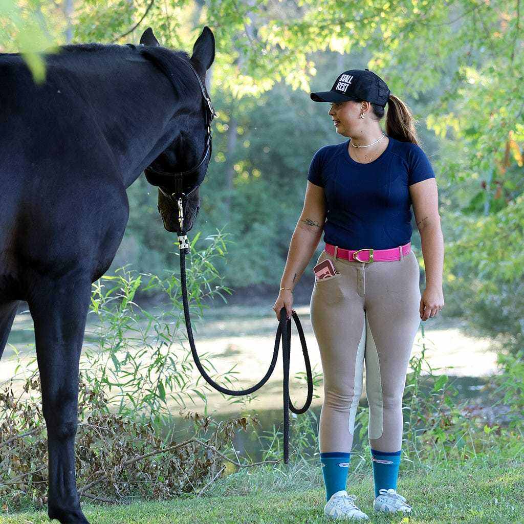 Woman wearing navy Equitation Tech Short Sleeve top with beige riding pants holding a black horse outdoors