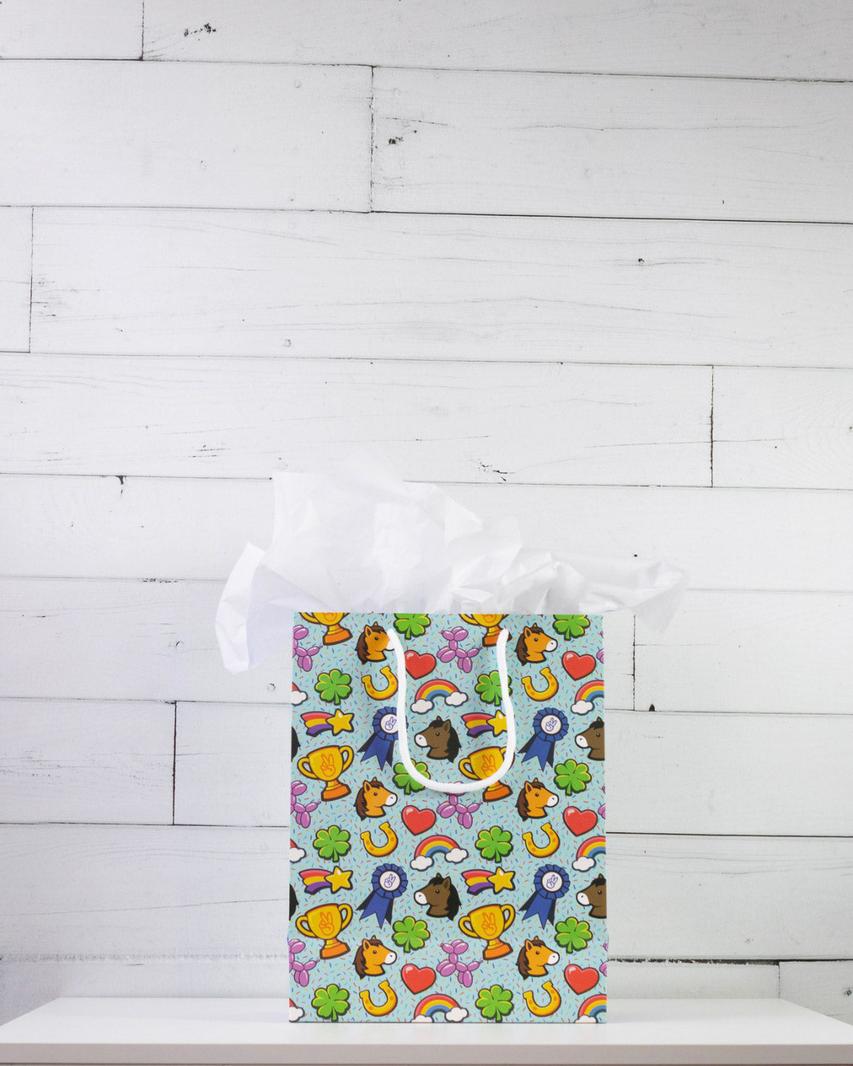 Full O' Luck medium gift bag on a white shelf, bright print of trophies, rosettes and four-leaf clovers.