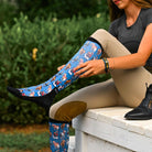 Blue holiday-themed boot socks with horse and peace sign patterns, flat seam toe, terry foot bottom, and stretchy black top worn by a person outdoors