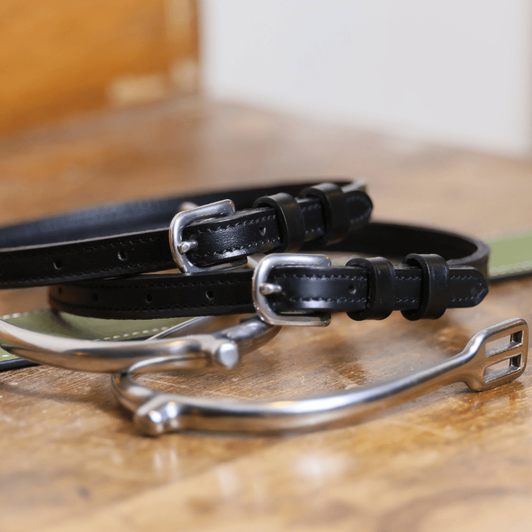 Close-up of black leather spur straps with stainless steel buckles and metal spurs on wooden surface from Not So Basic Black equestrian gift bundle