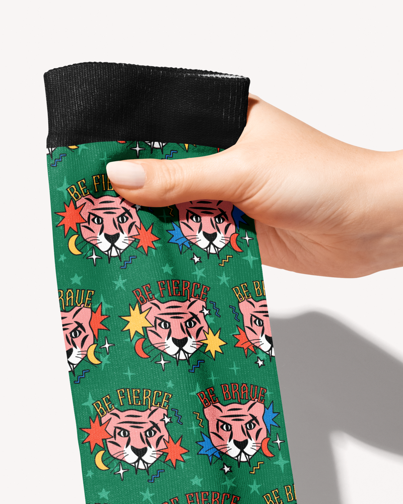 Hand holding the black cuff of the Be Brave boot sock, close-up of the pink tigers and Be Fierce lettering on green