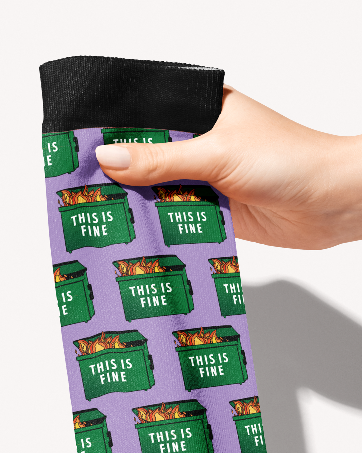 Close-up of the Dumpster Fire boot sock held by its black cuff, flaming green dumpsters reading This Is Fine on lilac
