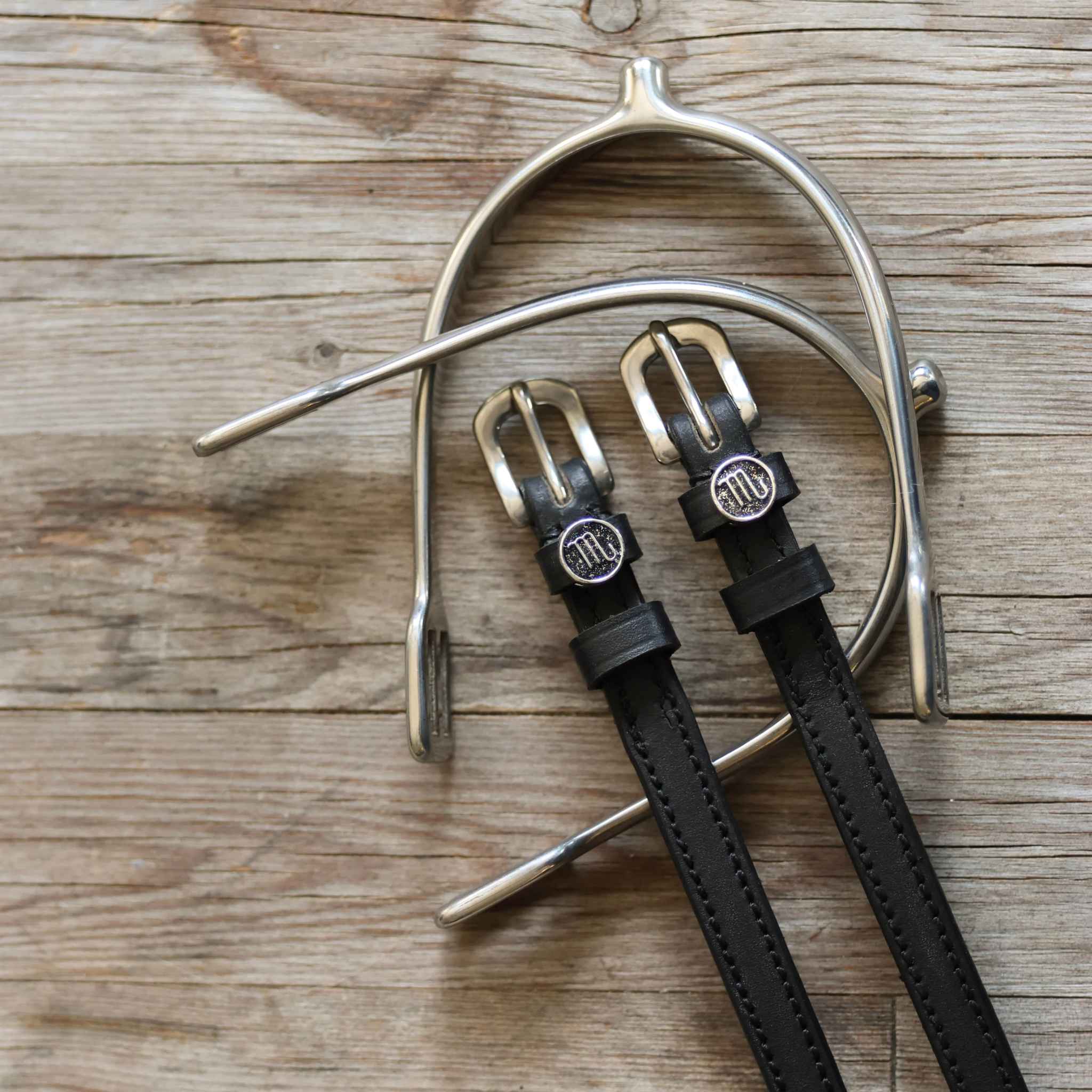 Black leather Scorpio Spur Straps with stainless steel buckles and custom charm on wood background