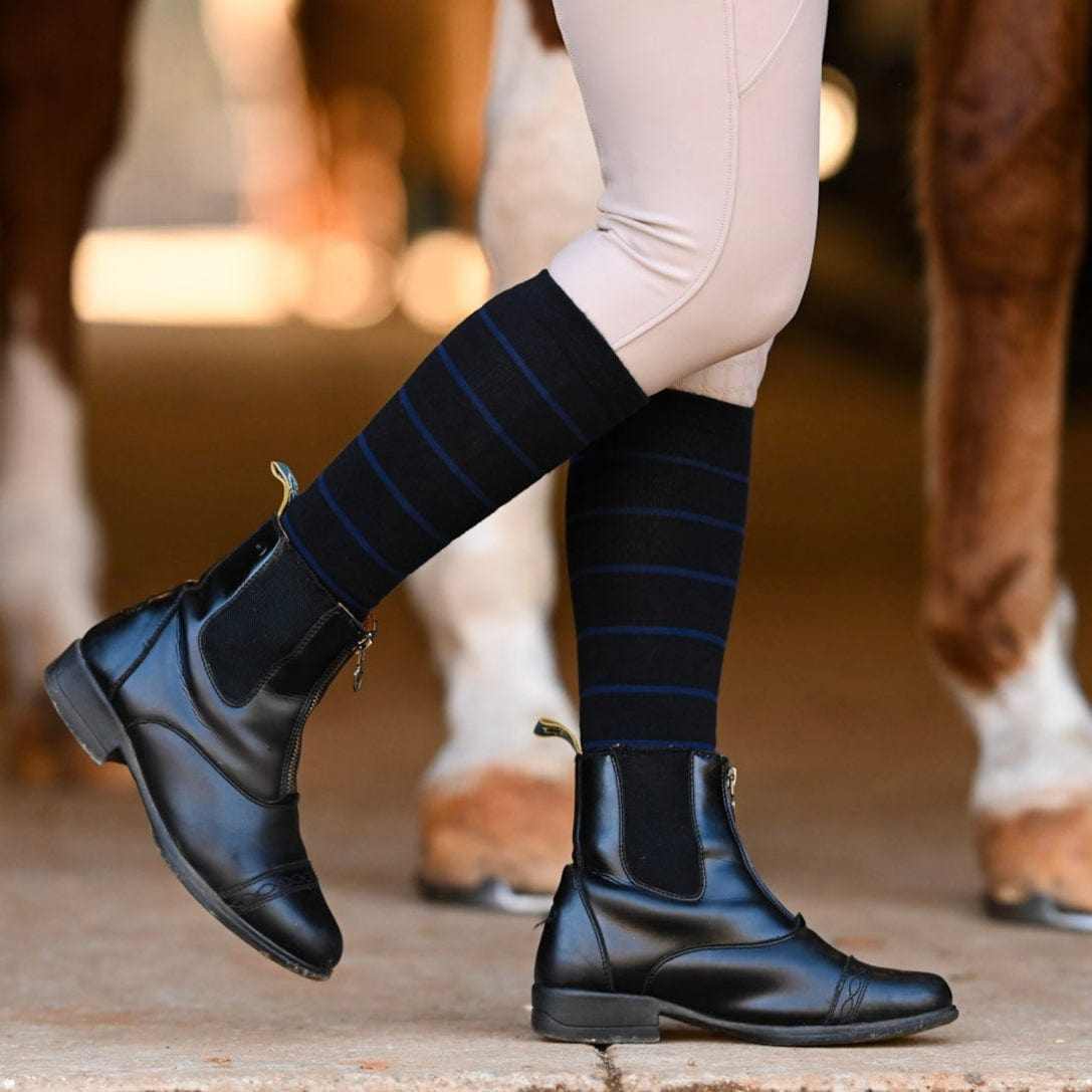 Close-up of person wearing black knit boot socks with blue stripes and black equestrian boots standing near horses