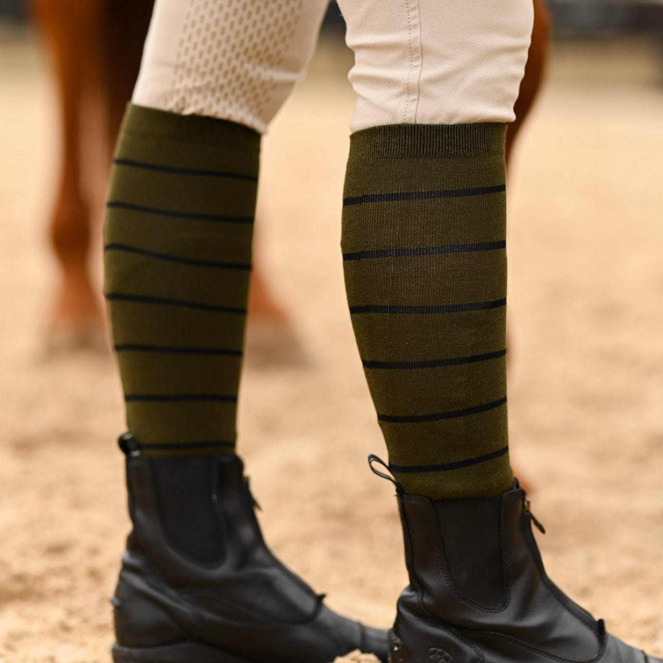 Close-up of olive green knit boot socks with thin black stripes worn with black riding boots and beige riding pants