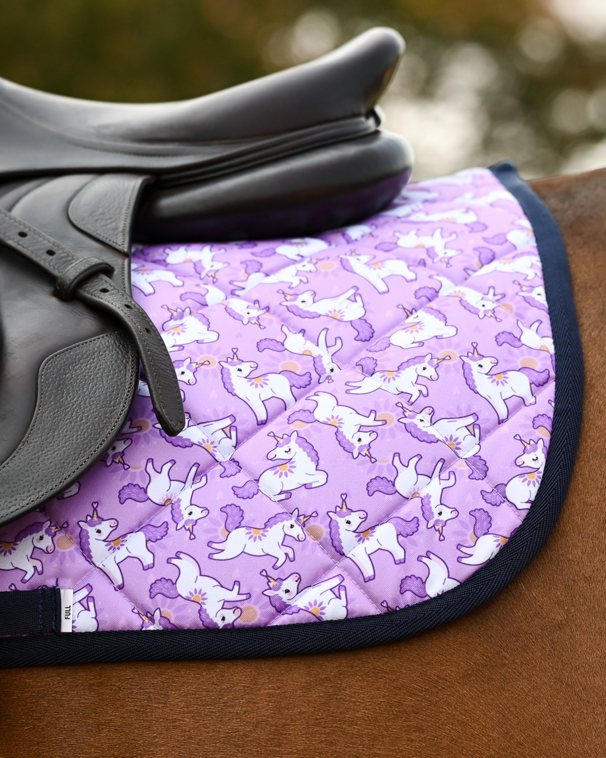Lilac unicorn-print saddle pad part-covered by a dark leather close-contact saddle on a bay horse