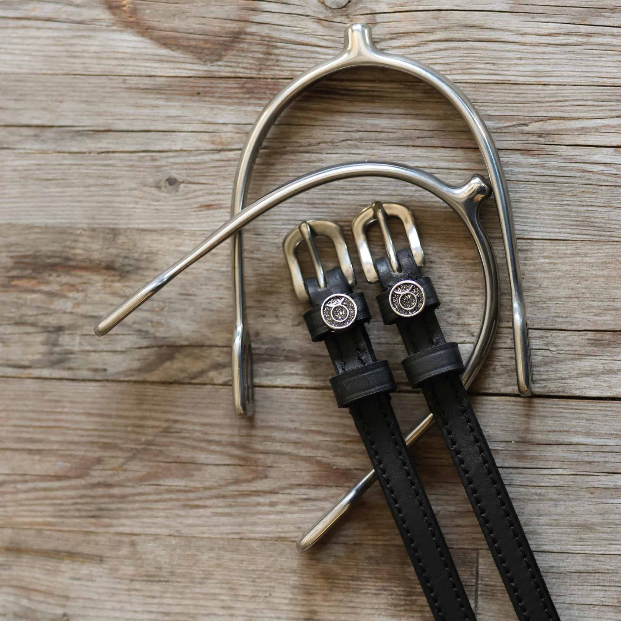 Taurus Spur Straps black leather with stainless steel buckles and custom charms on wooden surface with metal spurs